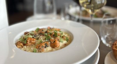 Valkoisessa kulhossa tarjoiltu kermainen risotto, jonka päällä paistettuja sieniä ja tuoreita yrttejä; taustalla punaviini‑ ja valkoviinilasi, vesikarahvi ja paahtoleipä erillisellä lautasella.