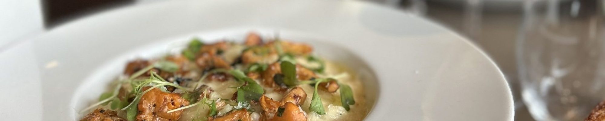 Valkoisessa kulhossa tarjoiltu kermainen risotto, jonka päällä paistettuja sieniä ja tuoreita yrttejä; taustalla punaviini‑ ja valkoviinilasi, vesikarahvi ja paahtoleipä erillisellä lautasella.
